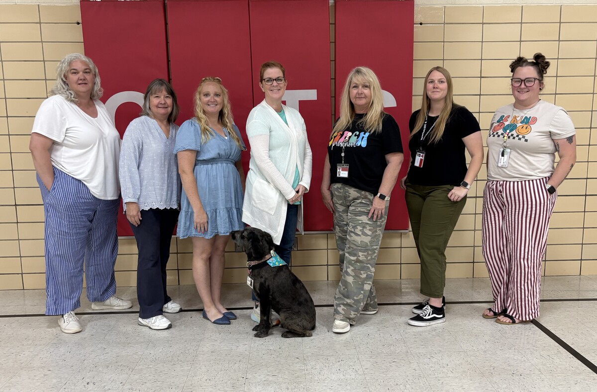 Preschool staff stanidng in a line in front of a red pad on a gym wall. A service dog is in front.